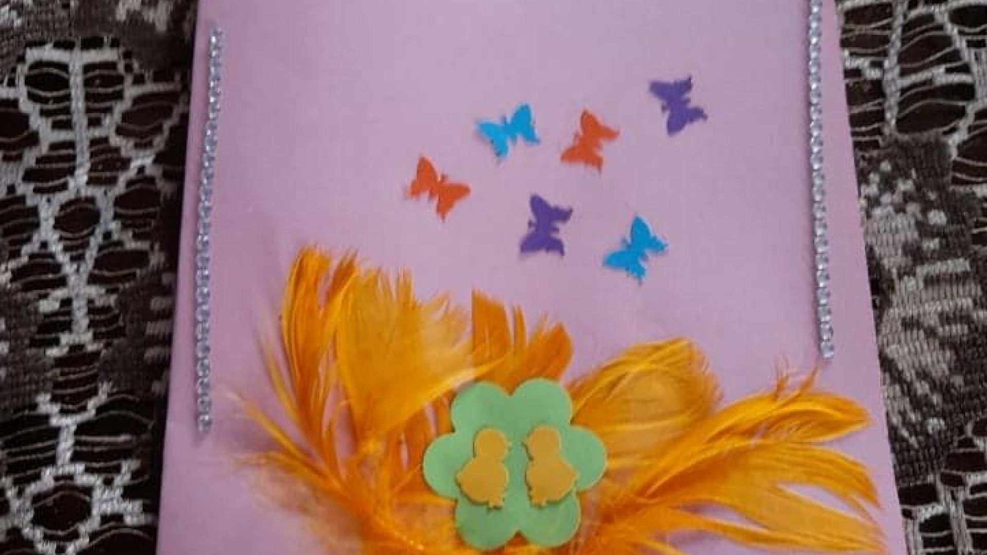 Kartka wielkanocna z żółtymi kurczaczkami przyklejonymi na zielonym kwiatku, z pomarańczowymi piórkami w tle i kolorowymi motylami. Autor: Agnieszka W. 