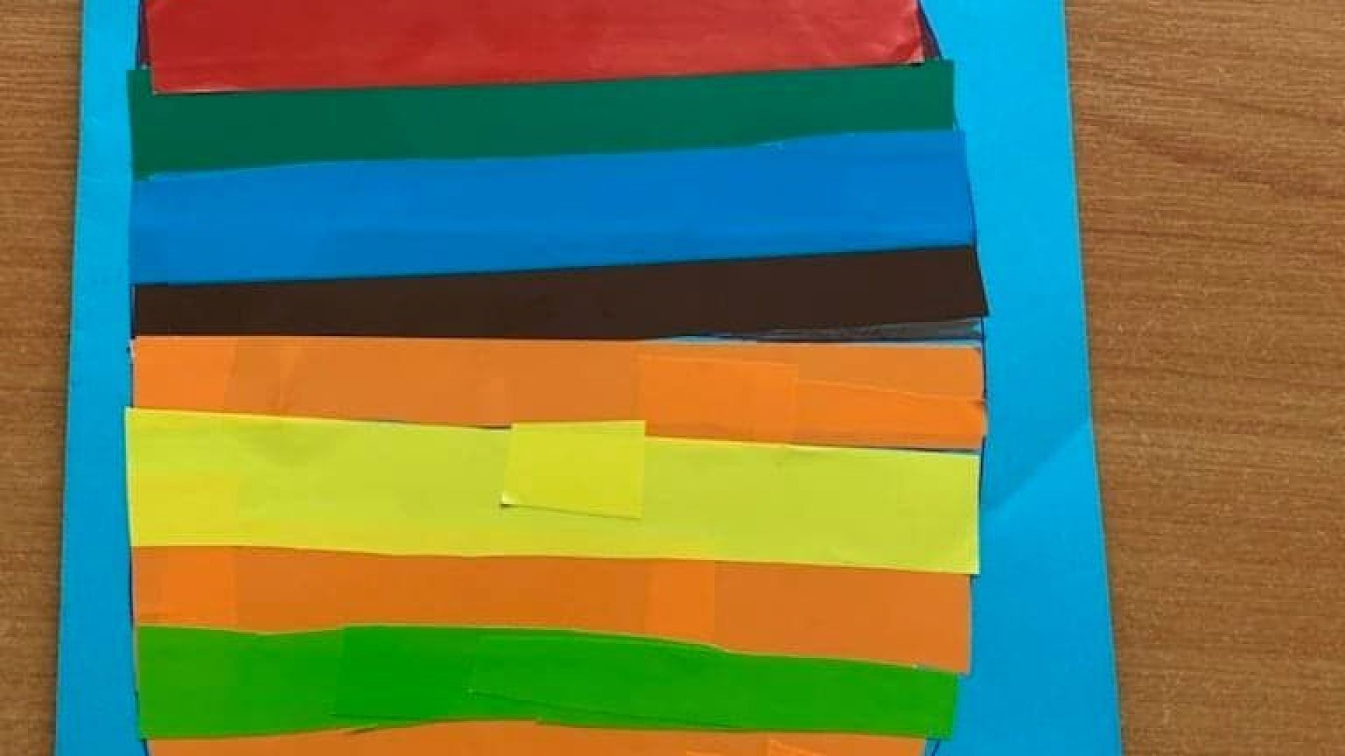 Wielkanocna kartka przedstawiająca dużą pisankę w kolorowe paski przyklejone z papieru kolorowego, na niebieskim tle, z napisem pisanka wielkanocna. Autor: Paweł K.