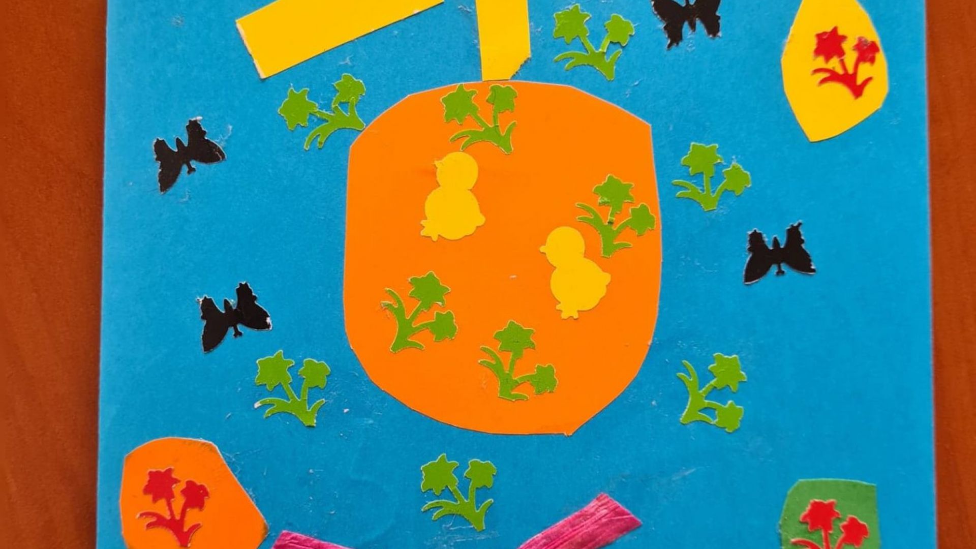 Kartka wielkanocna z zieloną trawą, trzema brązowymi zajączkami, kolorowymi kwiatami, motylami, pomarańczową pisanką z dwoma żółtymi kurczaczkami i czterema zielonymi kwiatami, z żółtym słońcem u góry i różową kokardą. Autor: Rafał T. 