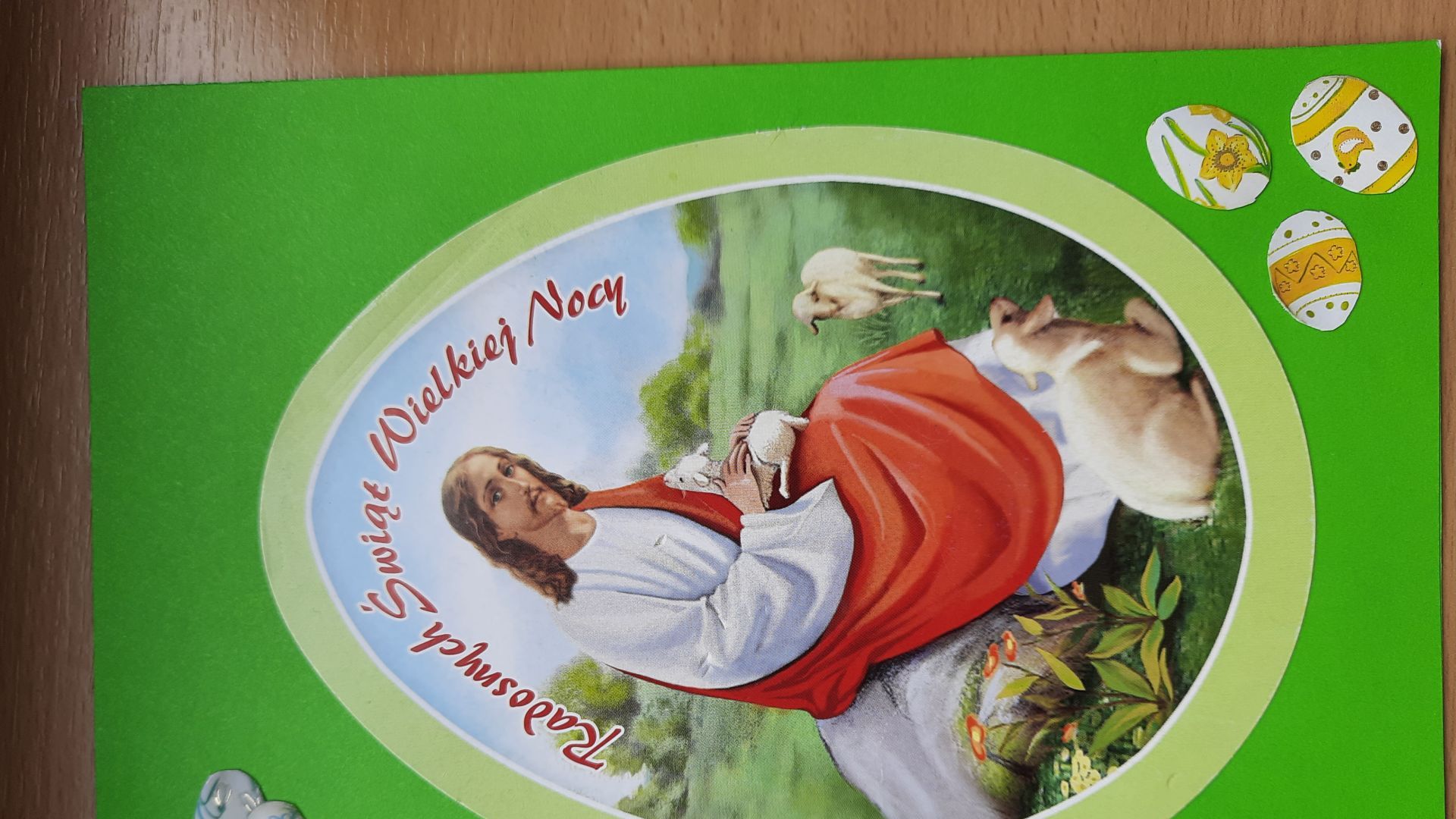 Kartka wielkanocna przedstawiająca Zmartwychwstałego Pana Jezusa z owieczkami na zielonym tle, z małymi pisankami po bokach. Autor: Sylwia W.