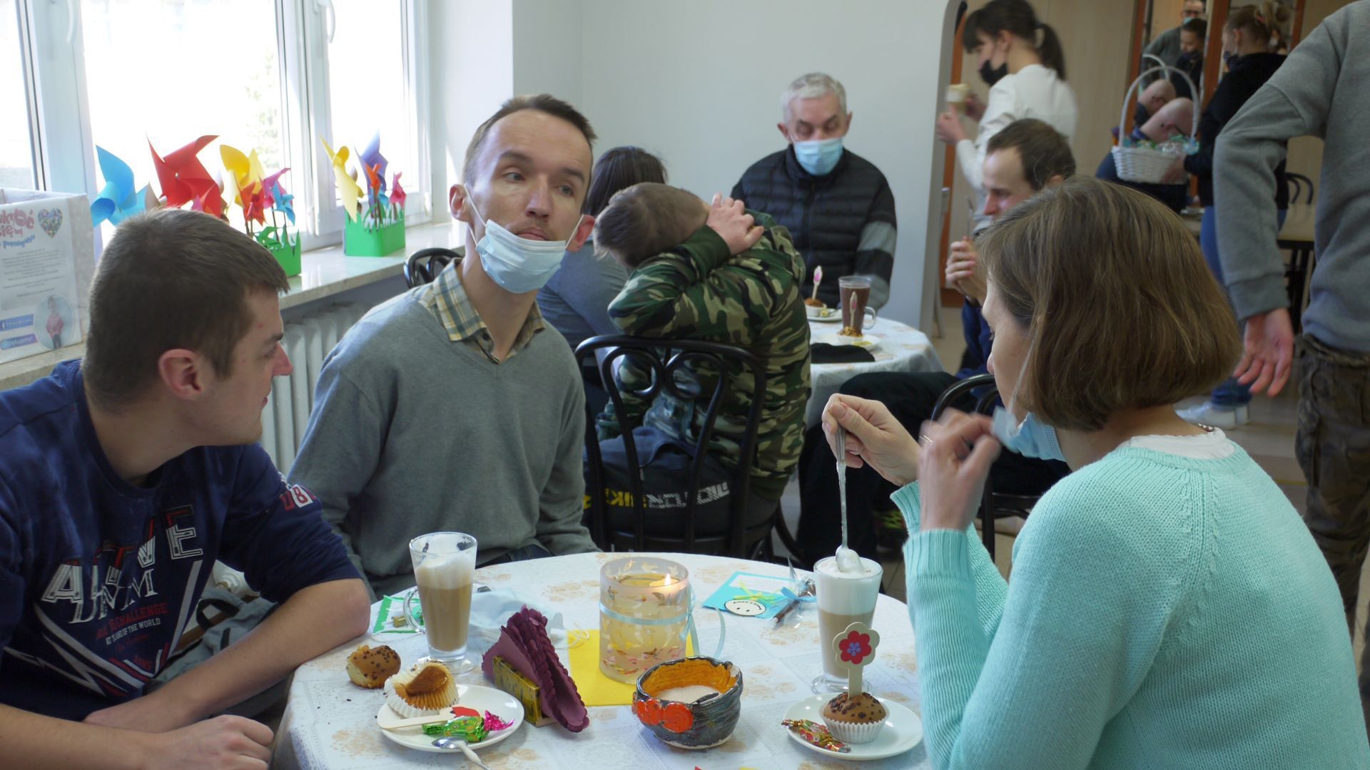 Uczestnicy oraz terapeuta siedzący przy stoliku w art.-kawiarni, na którym stoją kawy latte i talerzyki z babeczkami. 