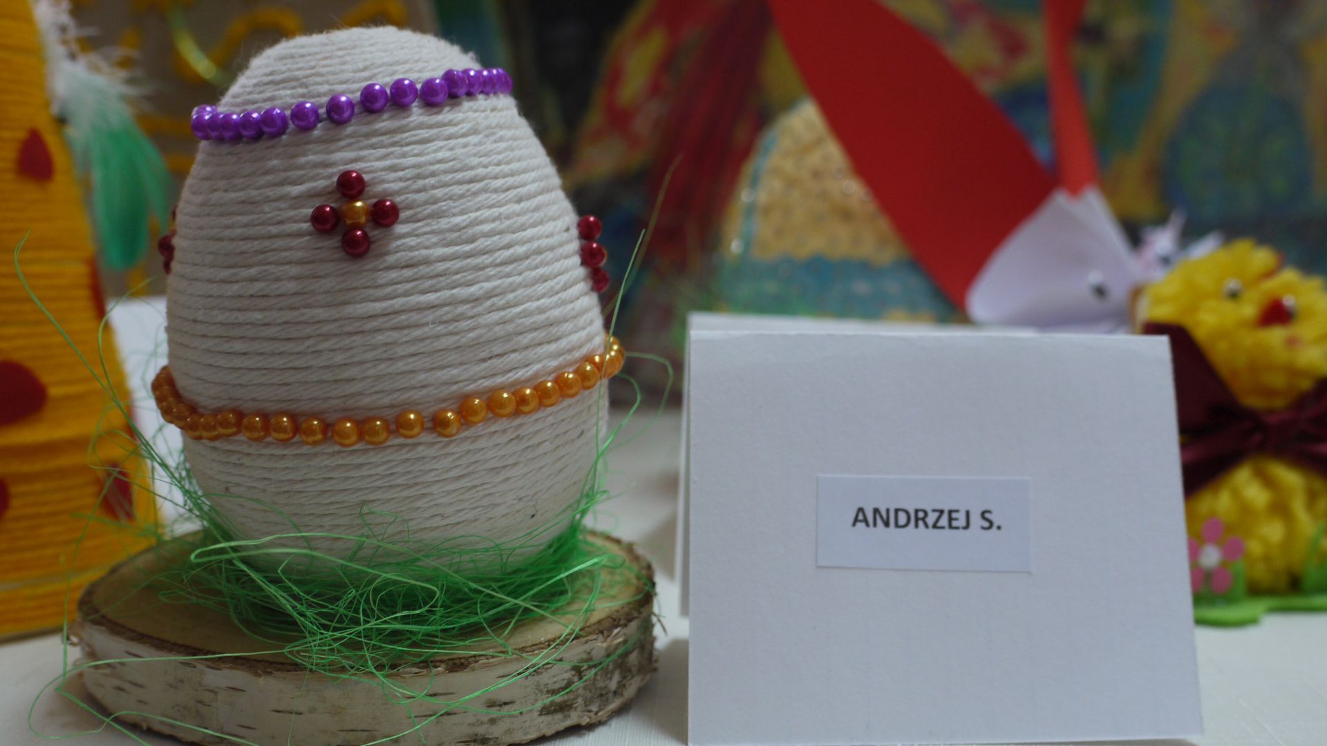 Pisanka wielkanocna owinięta jutowym sznurkiem ozdobiona pomarańczowymi i fioletowymi koralikami. Pisanka stoi na drewnianej podstawie - III miejsce w konkursie, autor Andrzej S.