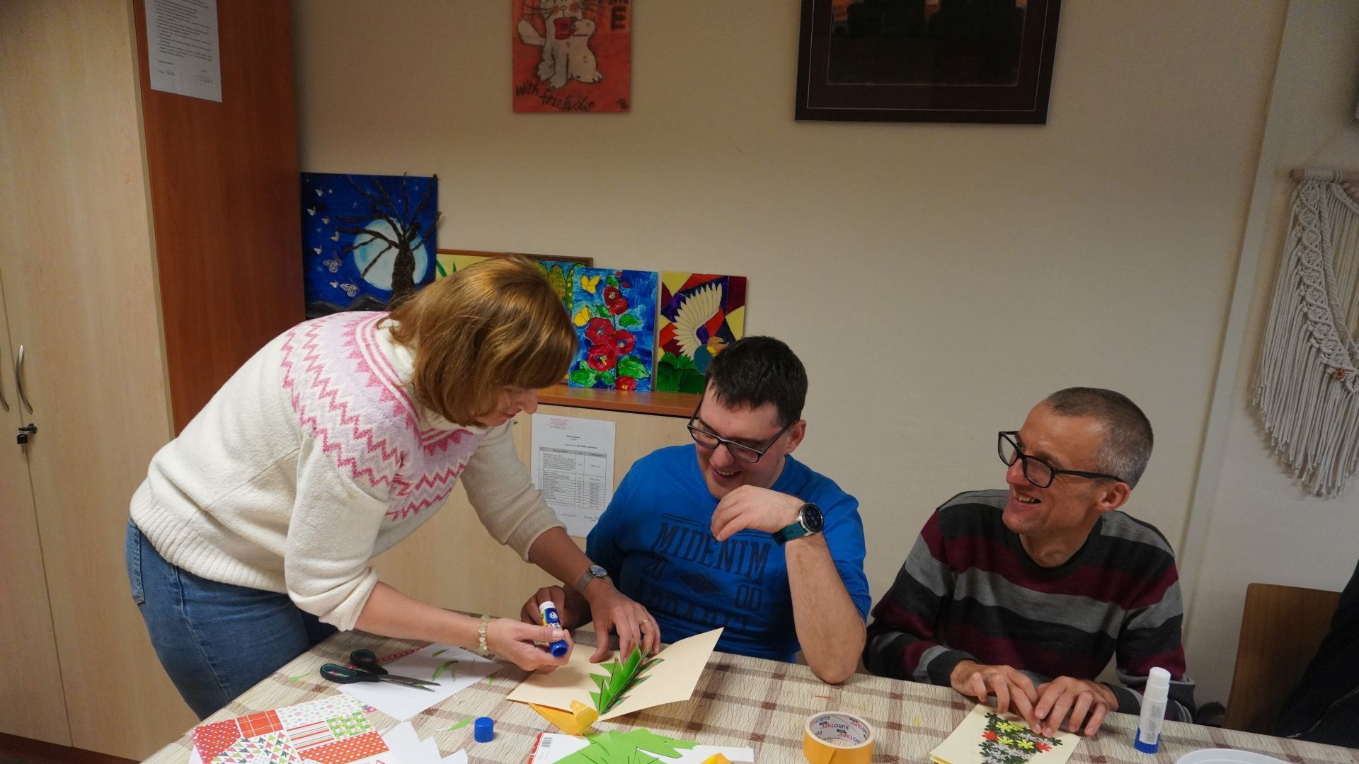 Terapeuta pomaga Uczestnikowi przykleić choinkę na kartkę świąteczną. 