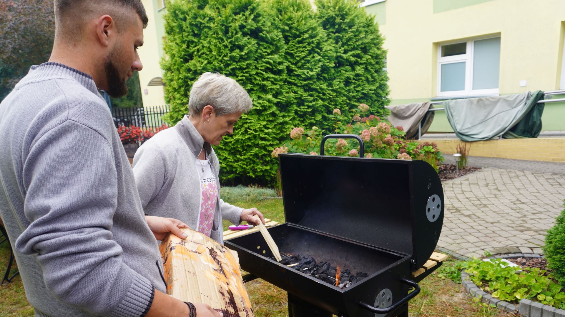 Uczestnik z opiekunem rozpala grilla w ogrodzie z okazji Dnia Pieczonego Ziemniaka. 