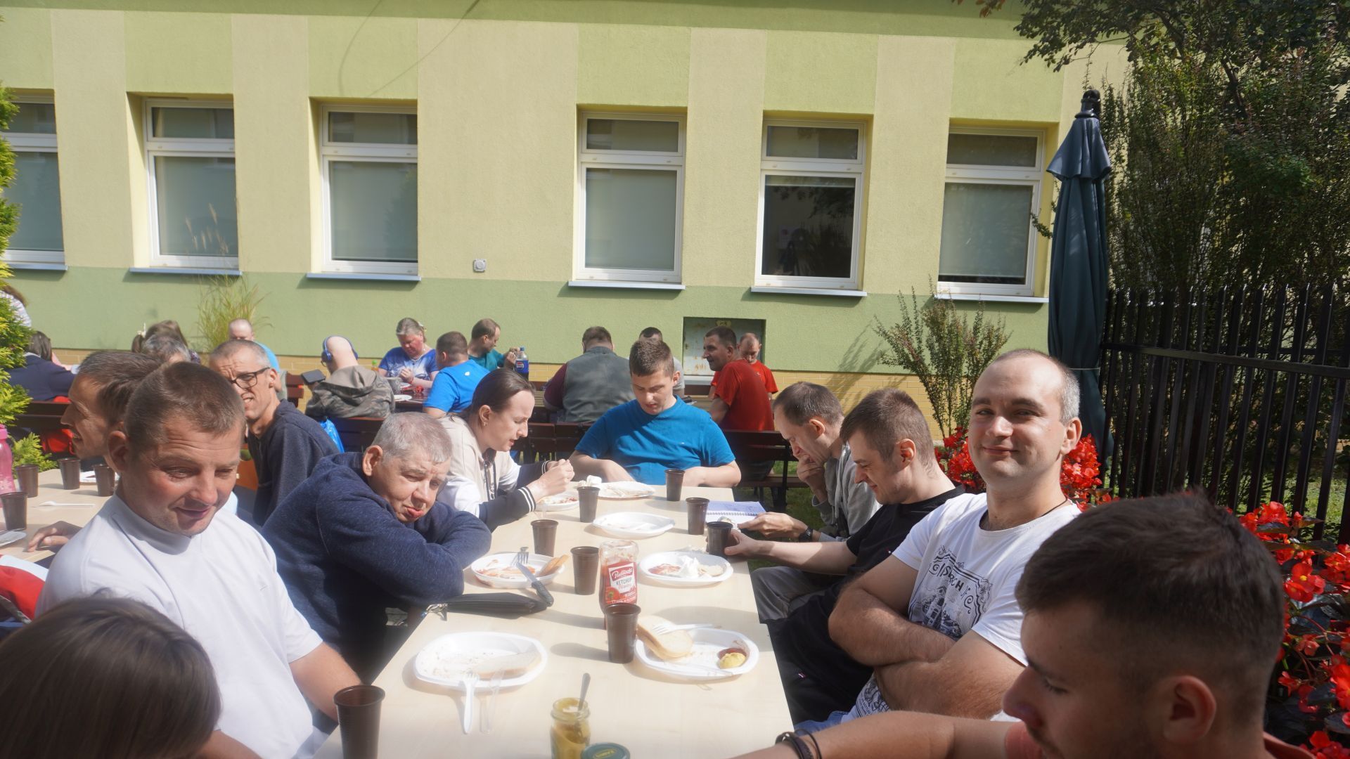 Uczestnicy siedzą za stołami podczas grilla w ogrodzie z okazji Dnia Pieczonego Ziemniaka. 