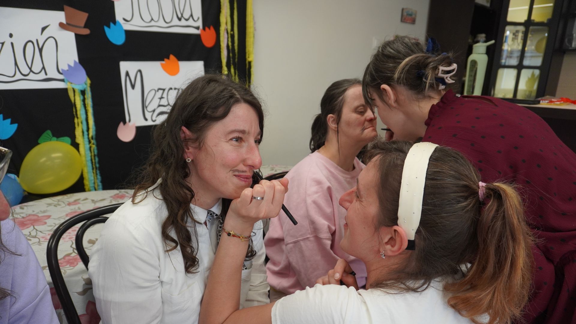 Uczestniczka Agnieszka W. maluje koleżance Justynie K. wąsy czarną kredką, podczas zajęć integracyjnych z okazji Dnia Kobiet i Mężczyzn. 
