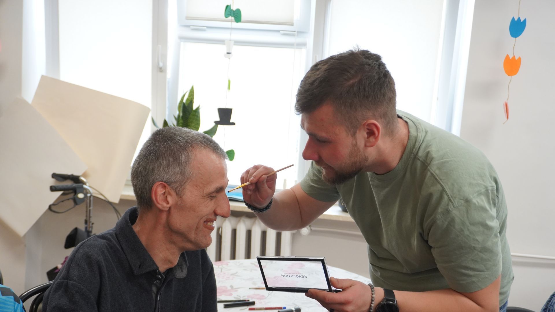 Uczestnik Bartłomiej wykonuje koledze Grzegorzowi makijaż podczas zajęć integracyjnych z okazji Dnia Kobiet i Mężczyzn.